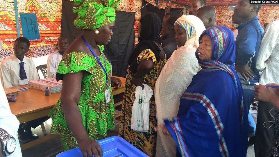 Chadian woman voting during the 2016 presidential election. Une Tchadienne se fait aider à placer son bulletin dans l'urne à côté d'autres électeurs qui attendent dans un bureau de vote à N'Djamena, Tchad, 10 avril 2016 (2016) Bagassi Koura (VOA) | Wikimedia