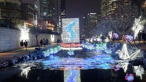 A Christmas festival at Cheonggyecheon displays two giant snowflakes ornaments on walls and a glowy and bright map of Korea surrounded by green butterflies with words displayed under the map stating “Merry Christmas, Peaceful Unification.”