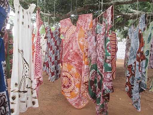 More than 10 African robes are hanging just above the ground by a thread from a sizeable African tree. The dresses are long in length but are all made of different and unique designs and colors. Some designs can be closely compared to a tye-dye style, while others have animals or distinct shape designs.